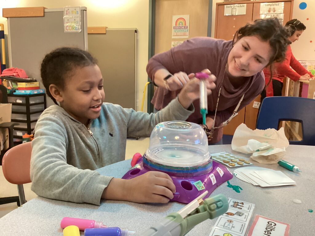 A student using a modified version of a spin art toy with AAC symbols and large buttons that are features of many adaptive play toys. 
