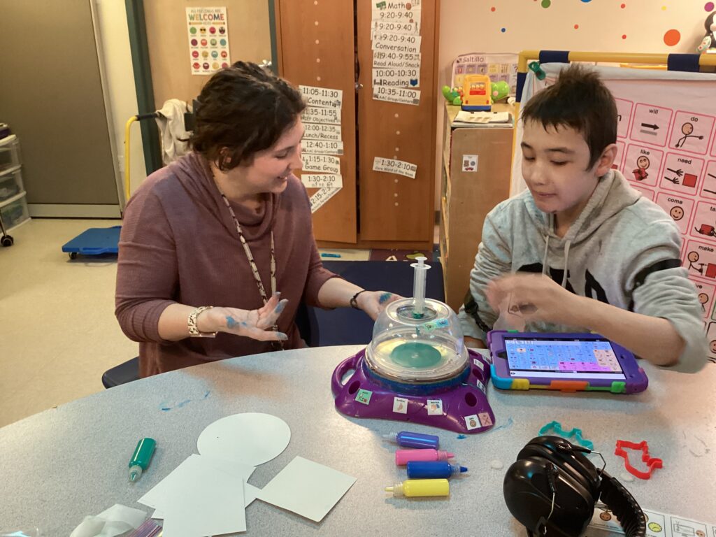 A student using a modified version of a spin art toy with AAC symbols and large buttons that are features of many adaptive play toys. 