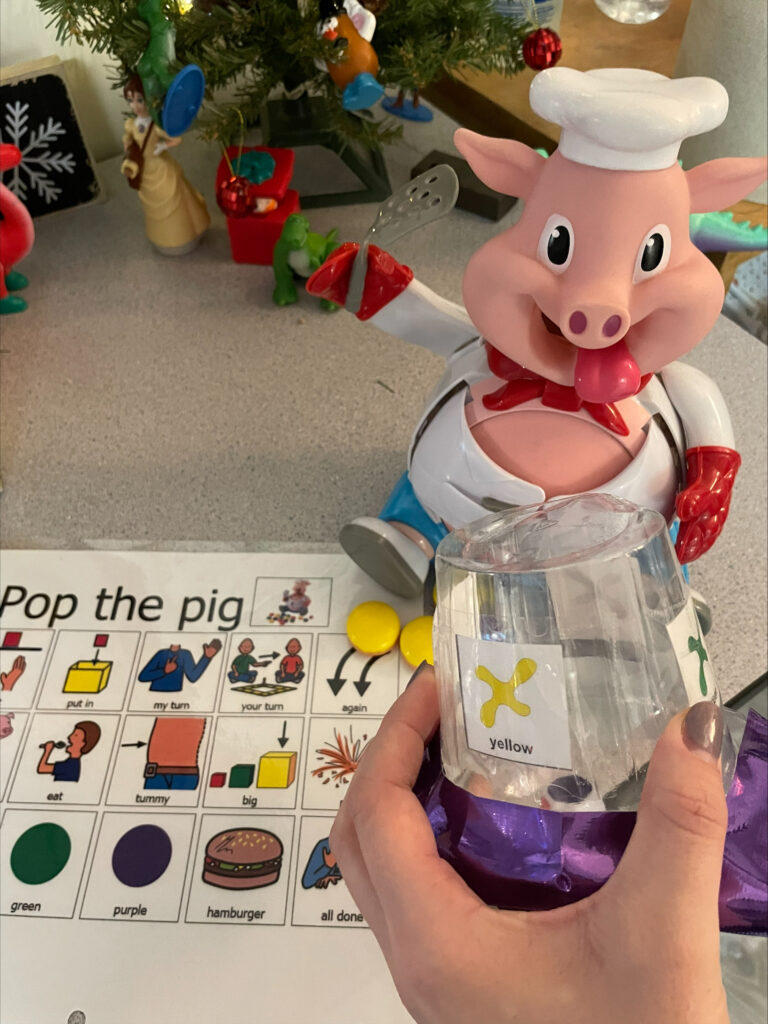 A student using a modified version of a Pop the Pig toy with AAC symbols, a container to shake dice, and large buttons that are features of many adaptive play toys. 